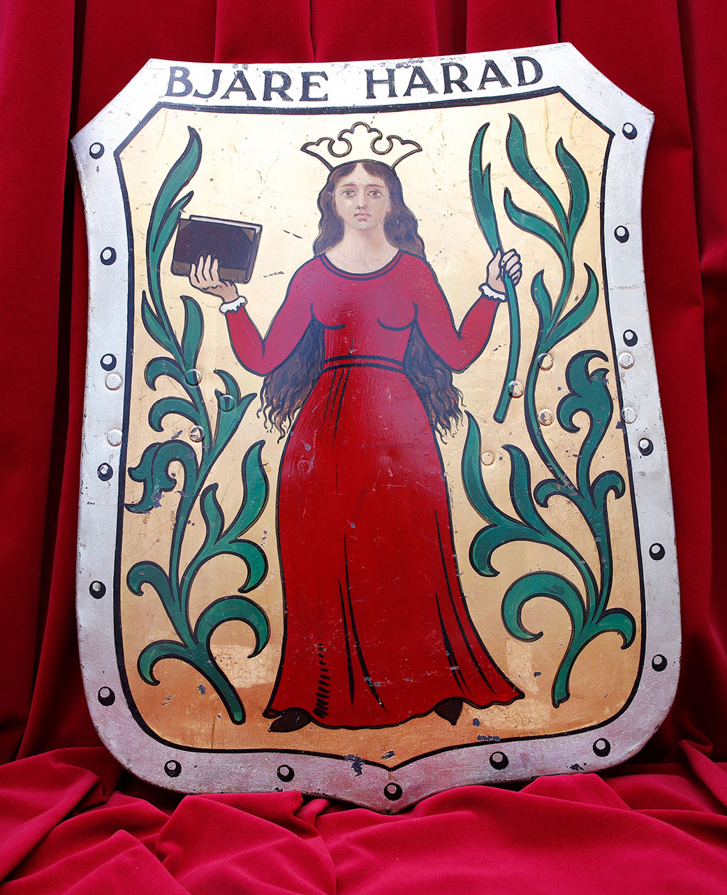 Guldig sköld med silverram. Kvinna i rött med krona, en bok och en palmkvist i vardera hand.